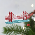 A holiday ornament featuring the Golden Gate Bridge in vibrant red and gold colors with a clear blue background, hanging from a Christmas tree.