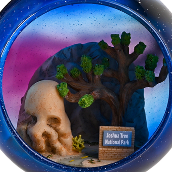 Glass christmas ornament of skull  rock and tree with a Joshua Tree National Park sign, set against a starry night sky zoomed in